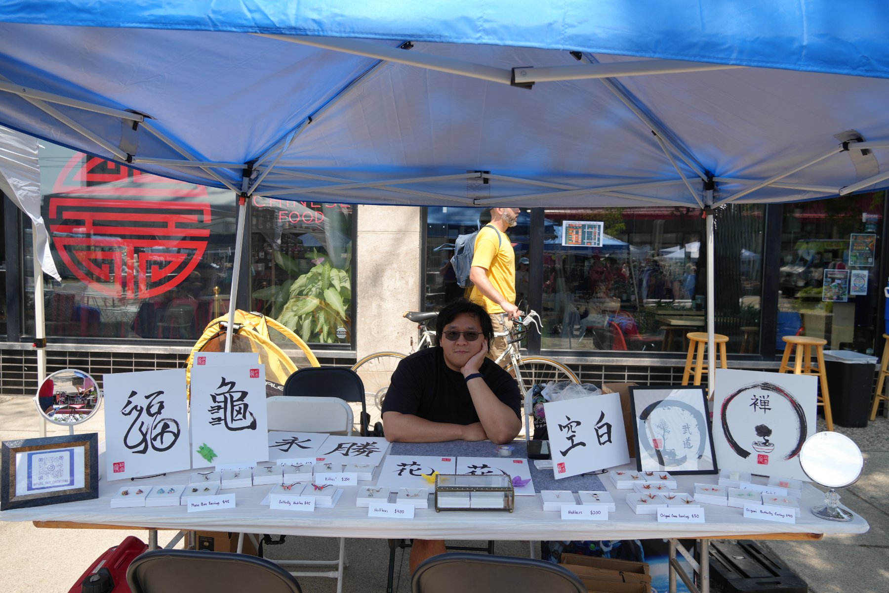 Skyler seated behind the Pure Sky Creations stall under a blue canopy, calligraphy boards and boxed jewelry across the table
