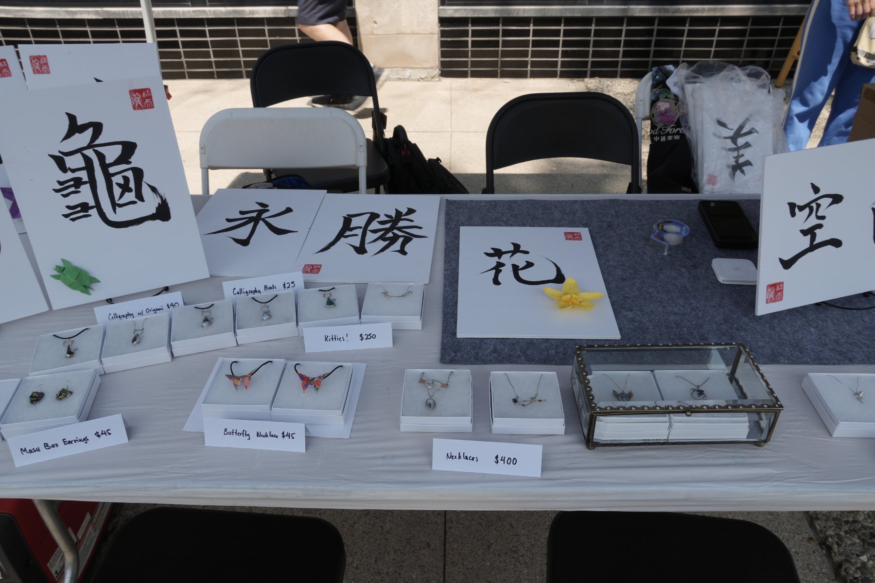 永, 勝 and 花 prints above origami butterfly necklaces, kitten pendants and silver necklaces in a glass case