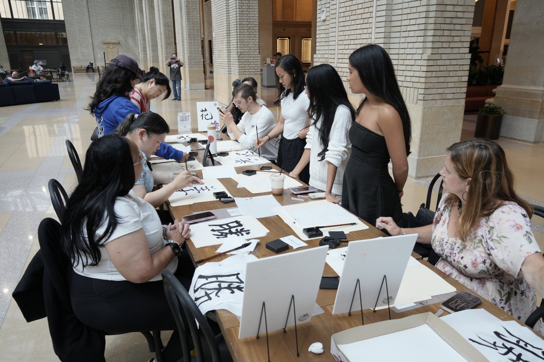 Students seated along a long table in the station's hall, brushing characters on white paper beside ink stones