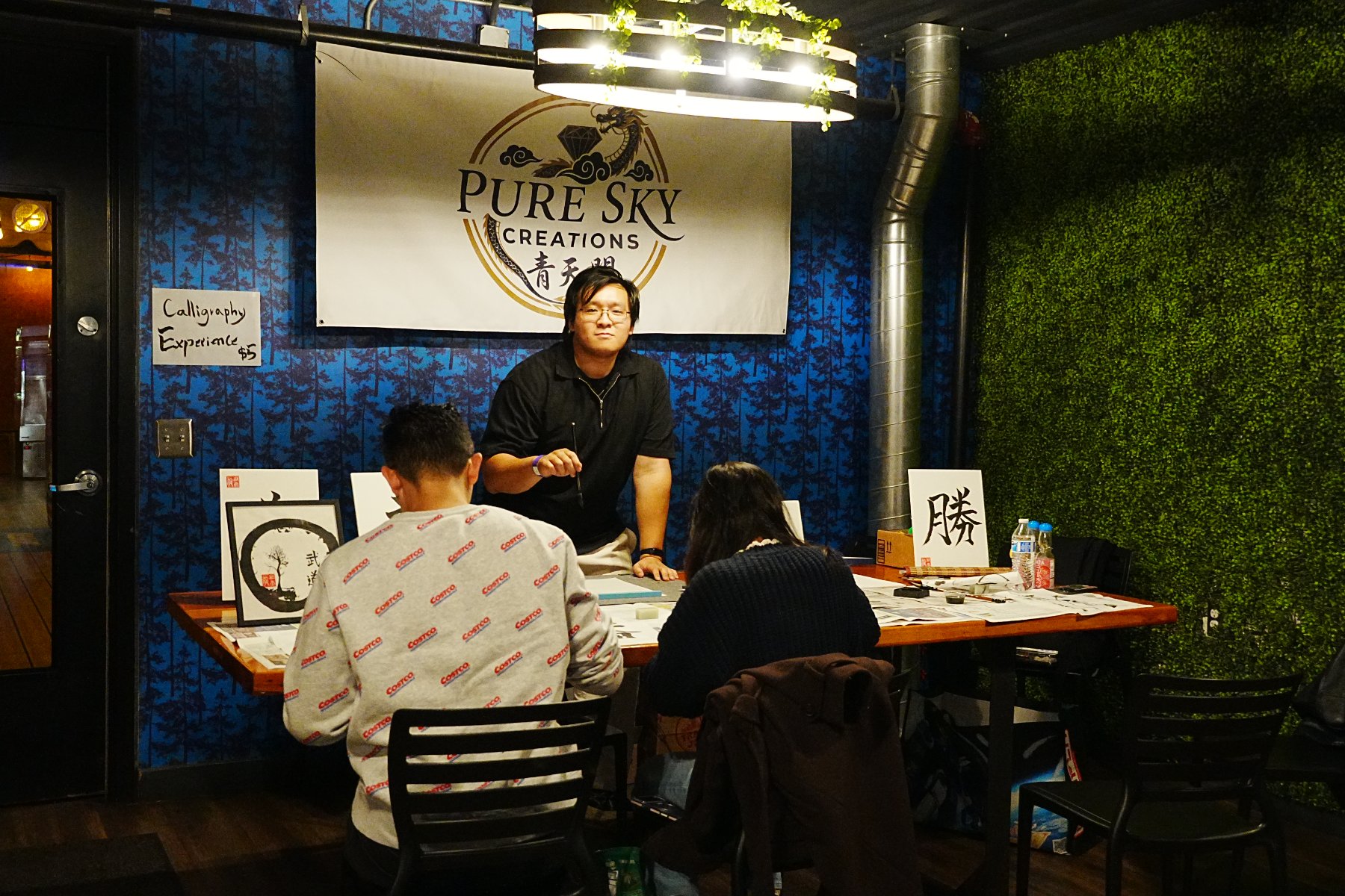Skyler guiding two guests at the calligraphy booth beneath the Pure Sky Creations banner, a 勝 board and an ensō on the table