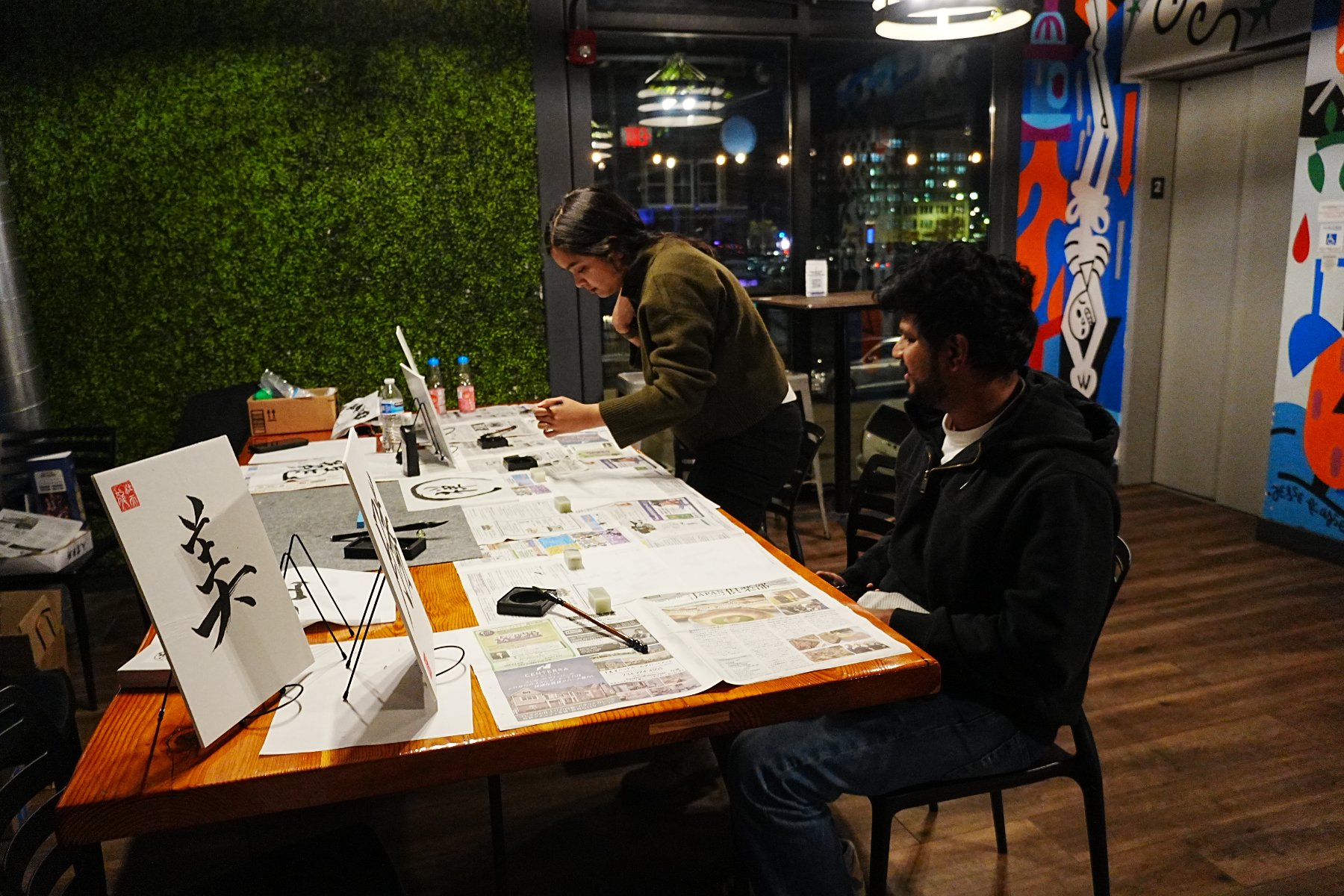 Two guests at work along the long table, a 楽 board on a stand in the foreground