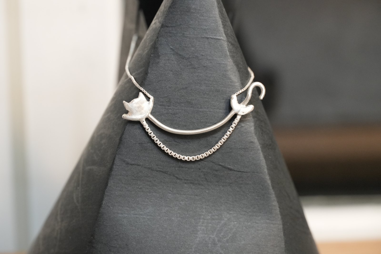 Jiggly Belly, front view: a silver cat head and curled tail joined by a curved silver bar, with a box chain draped beneath like a belly