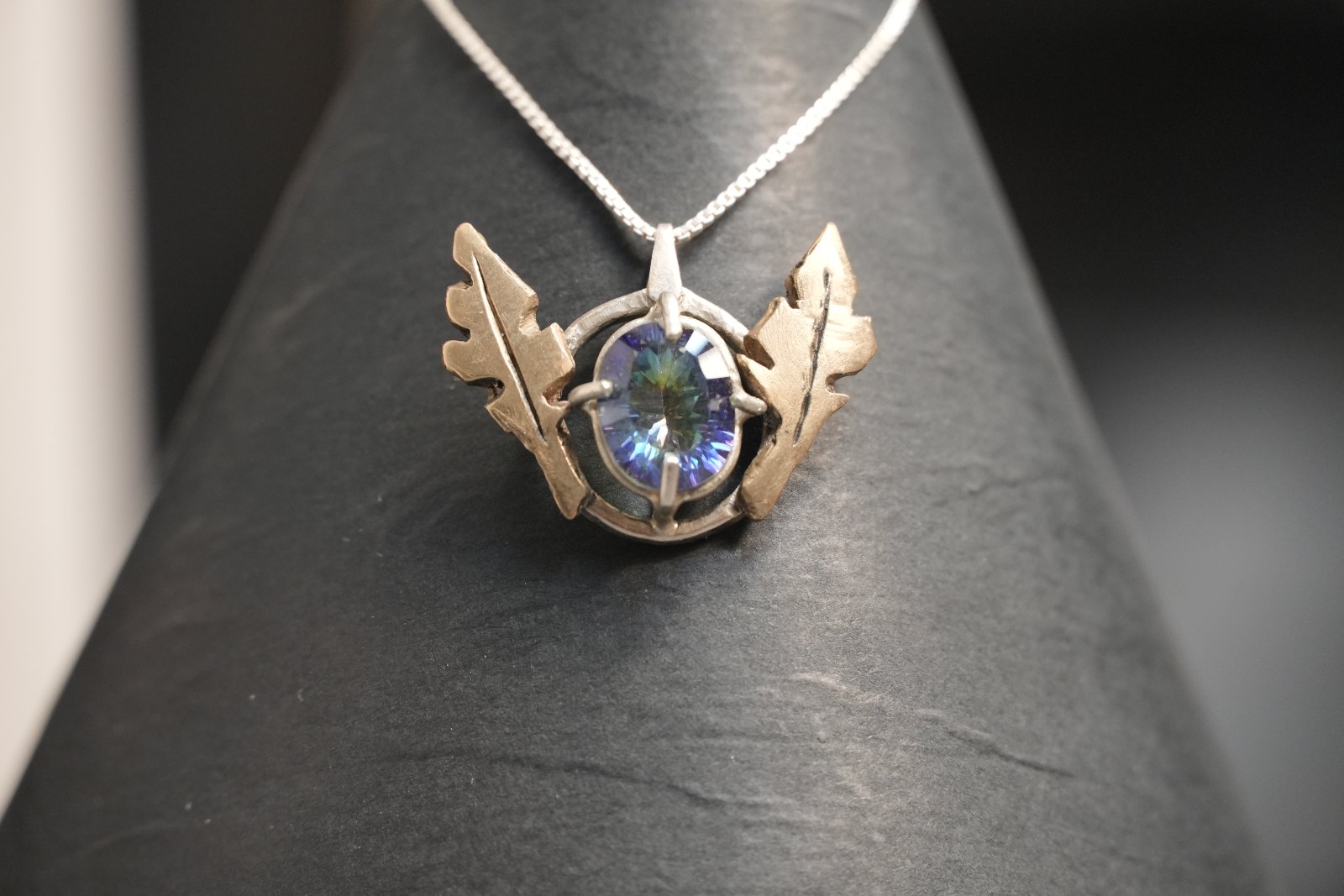 Mystic Topaz Pendant, front view: oval mystic topaz prong-set in a sterling silver ring between two brass oak-leaf wings, on a box chain