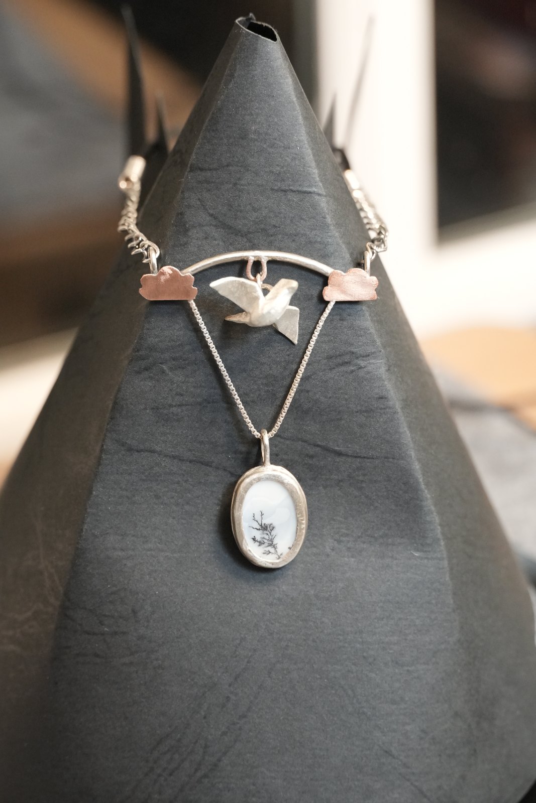 Sylvan, front view: a silver bird in flight hangs from an arched bar between two copper clouds, above an oval dendritic-opal pendant showing a branching tree