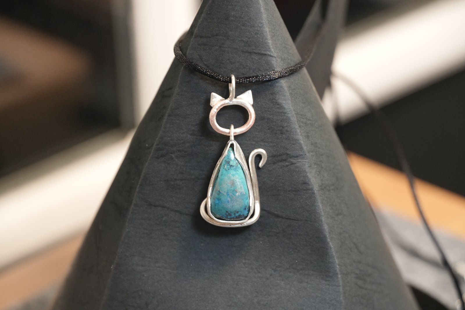 Teardrop Kitten in African azurite, front view: silver cat with pointed ears and curled tail framing a teal-and-indigo teardrop, on a black cord