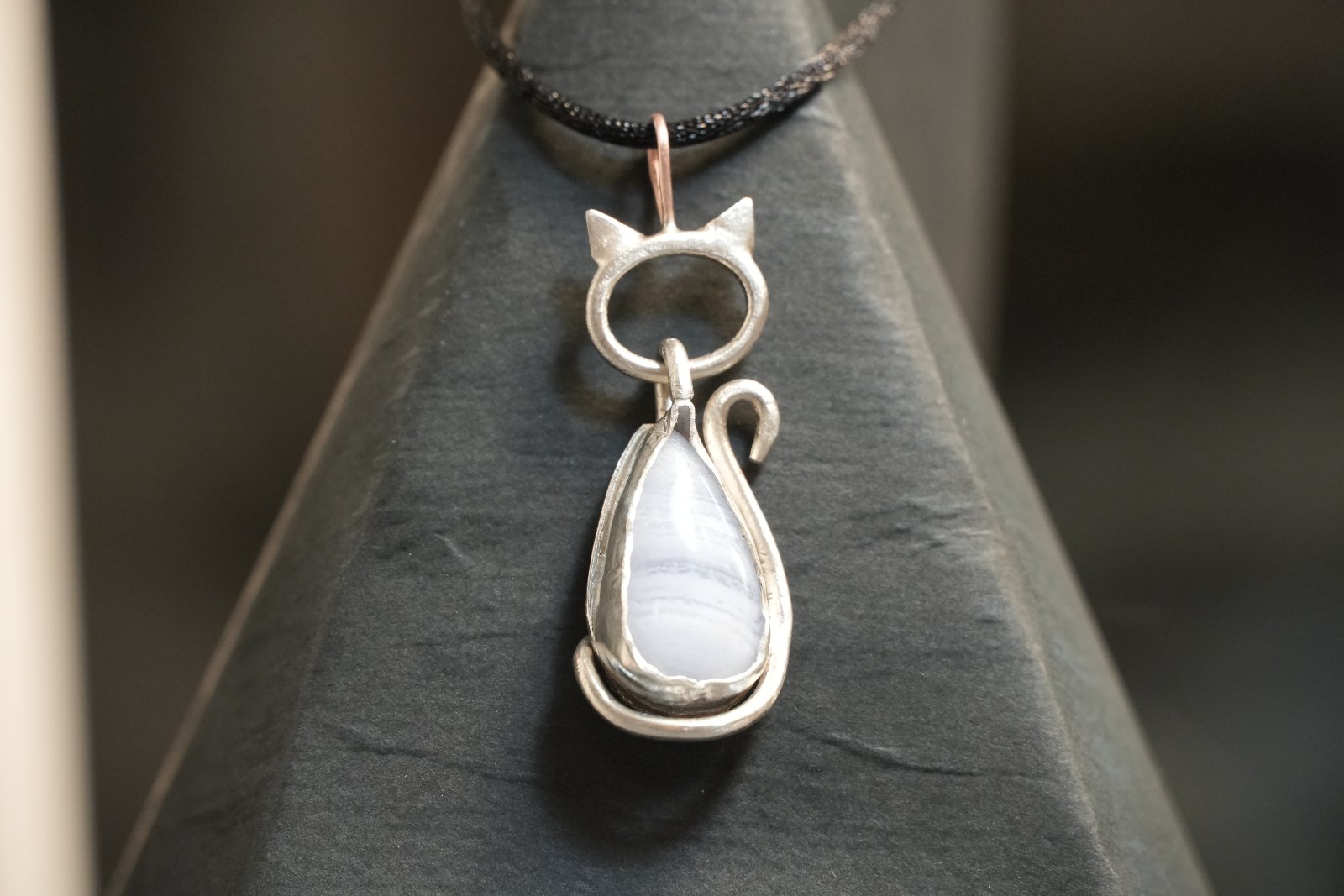 Teardrop Kitten in blue lace agate, front view: silver cat with pointed ears and curled tail framing a pale banded blue teardrop, on a black cord