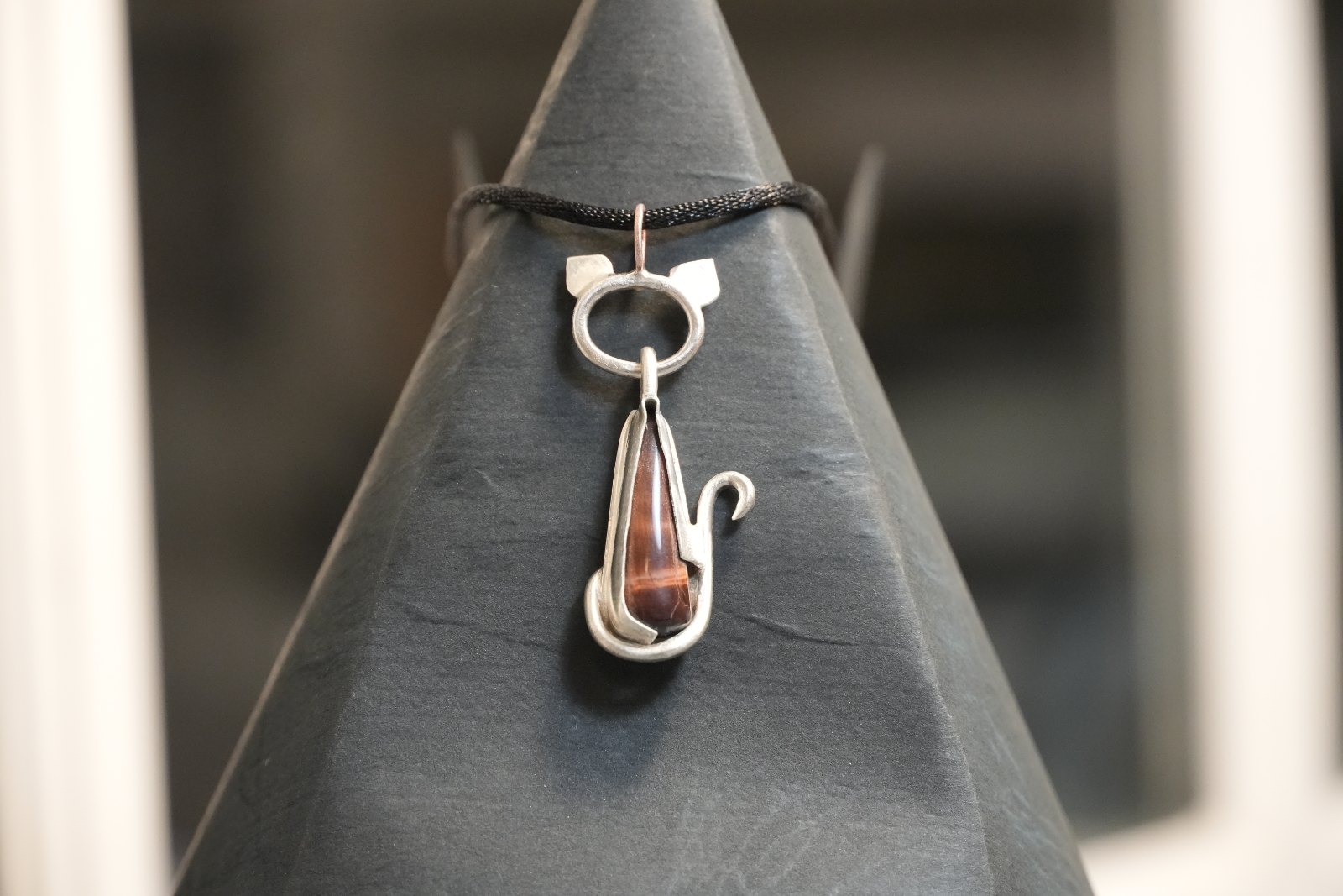 Teardrop Kitten in red tiger eye, front view: silver cat with pointed ears and curled tail framing a slender red-brown chatoyant teardrop, on a black cord