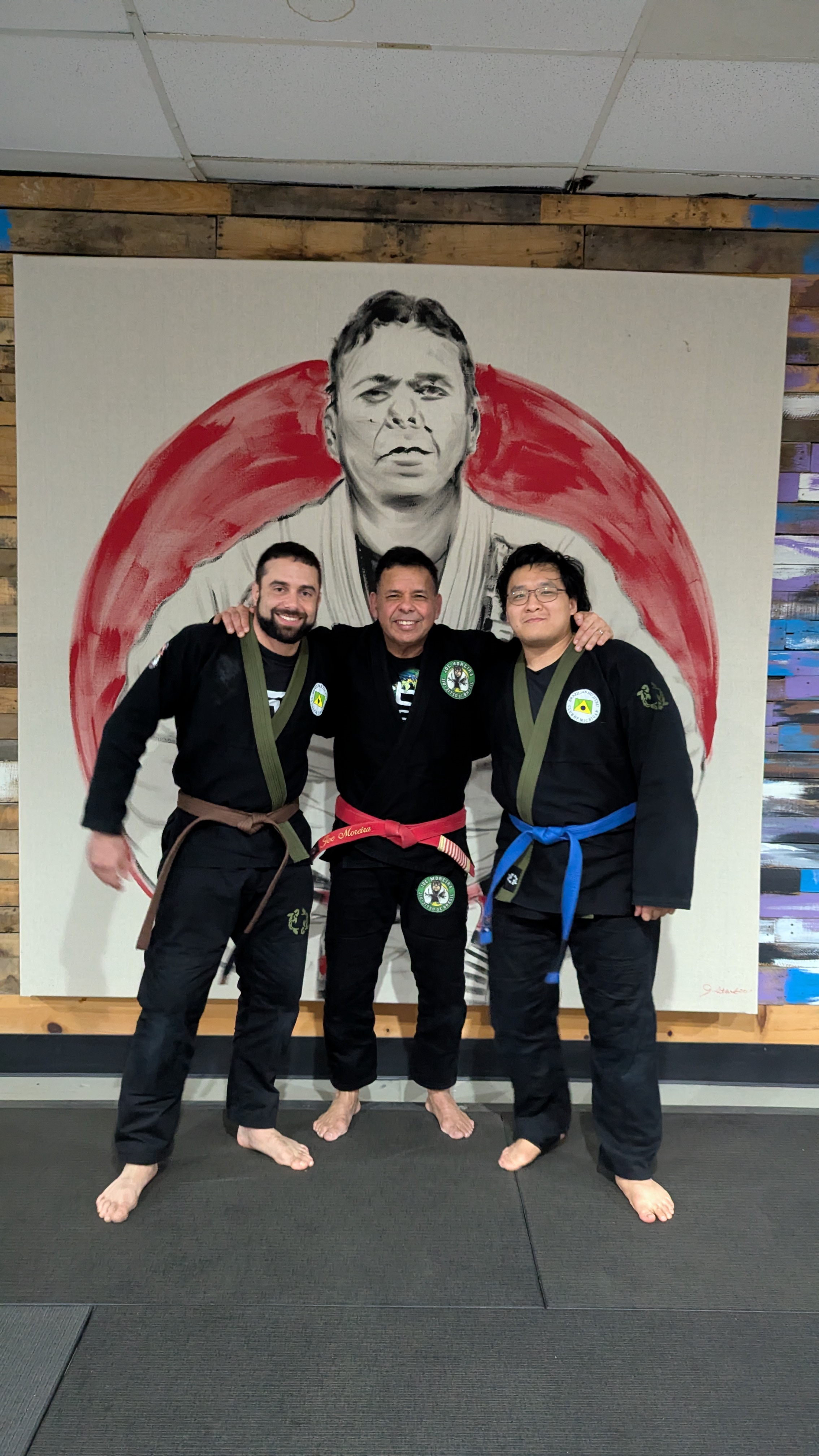 Three Brazilian Jiu-Jitsu practitioners in gi with colored belts before a painted portrait