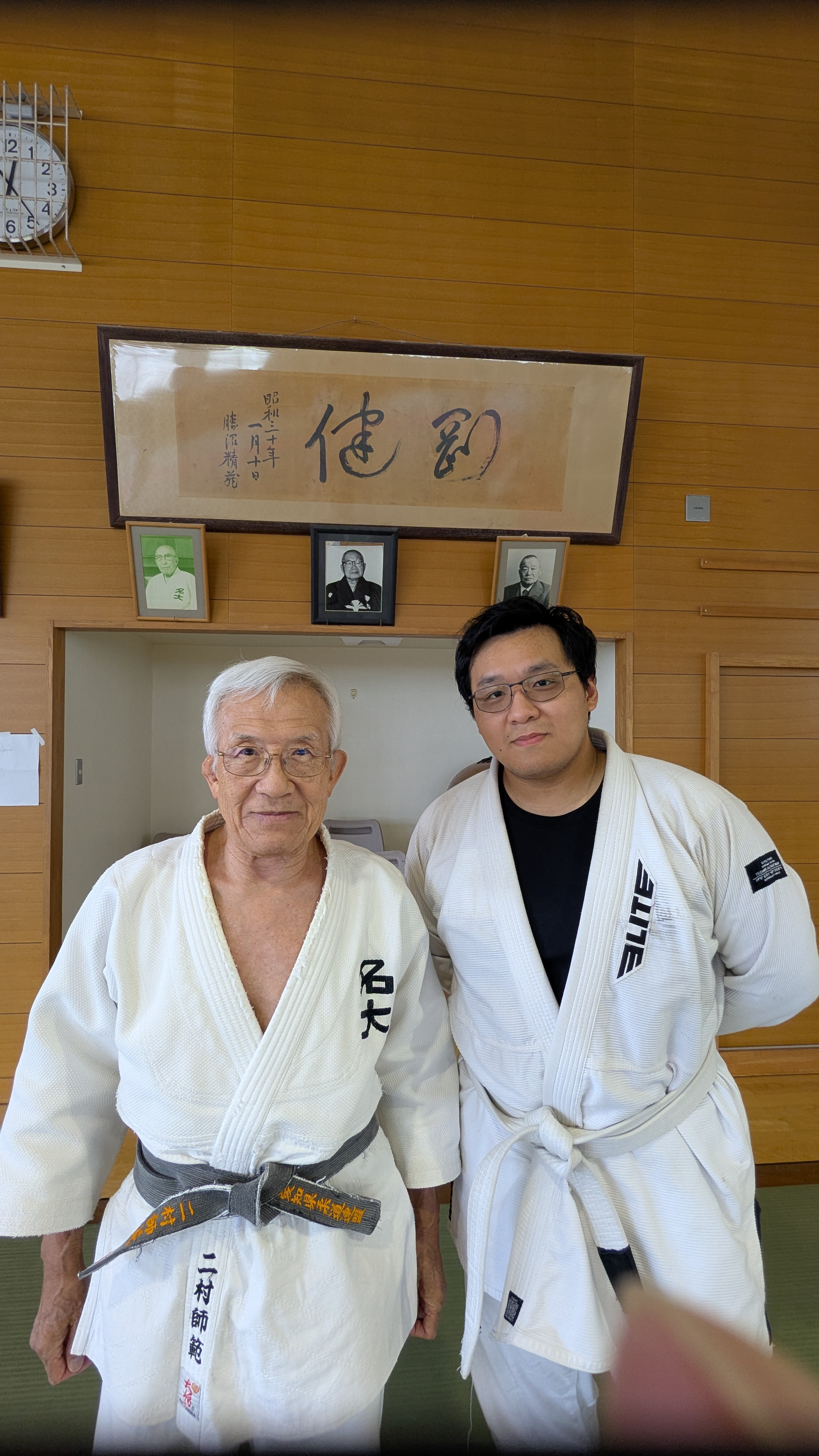 Judo practitioner with an elder sensei beneath framed calligraphy and portraits