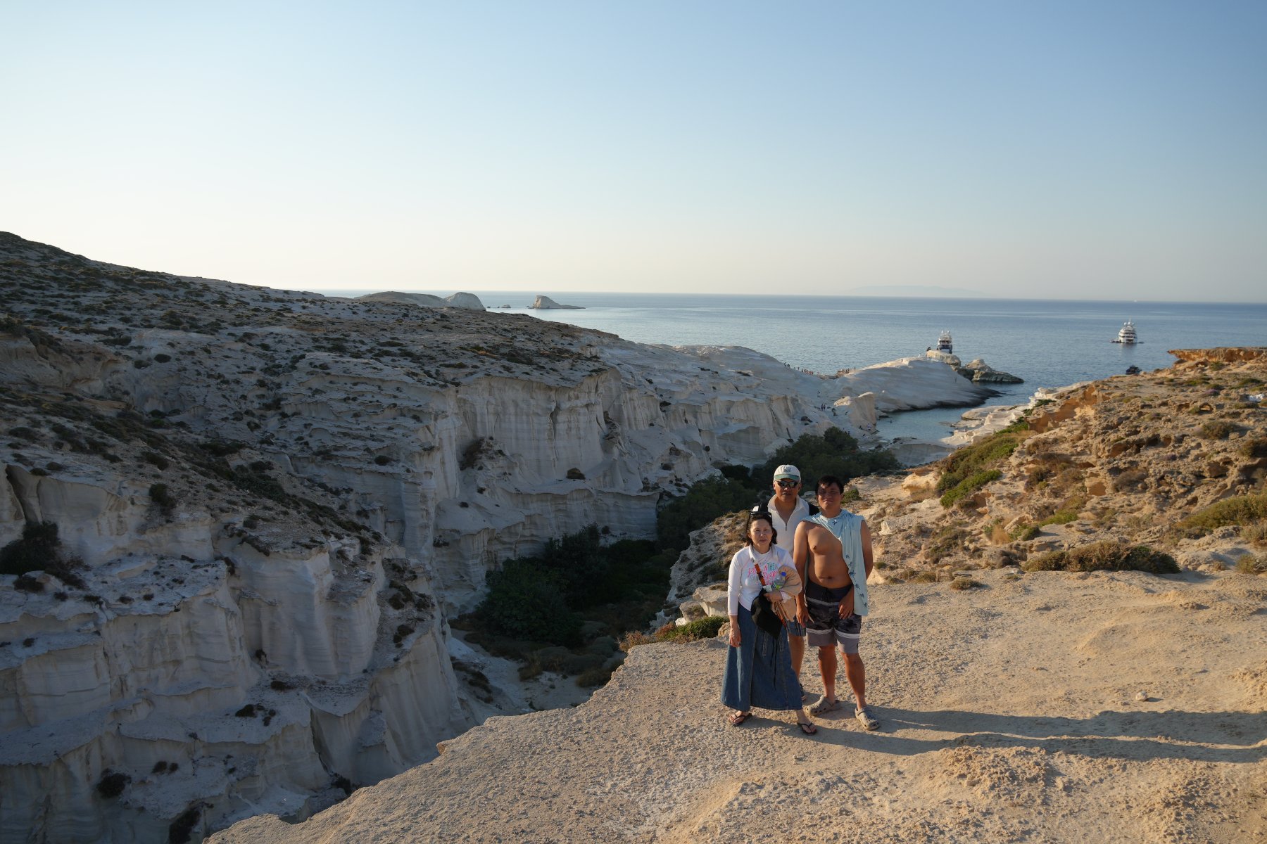 A family standing on a sandstone ledge above the white volcanic canyons of Sarakiniko, the Aegean behind