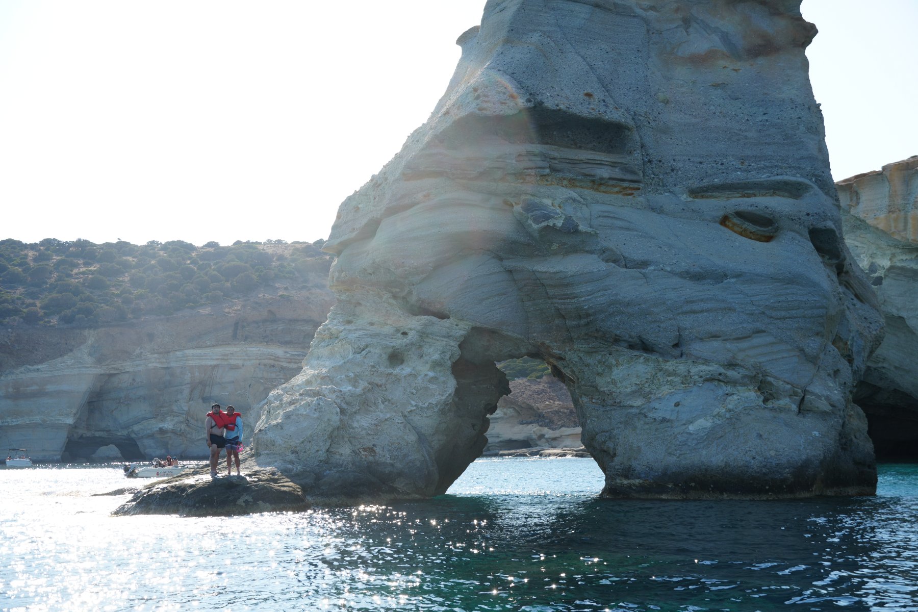 A towering sea-carved rock arch over glittering turquoise water, two swimmers in red life vests on a ledge
