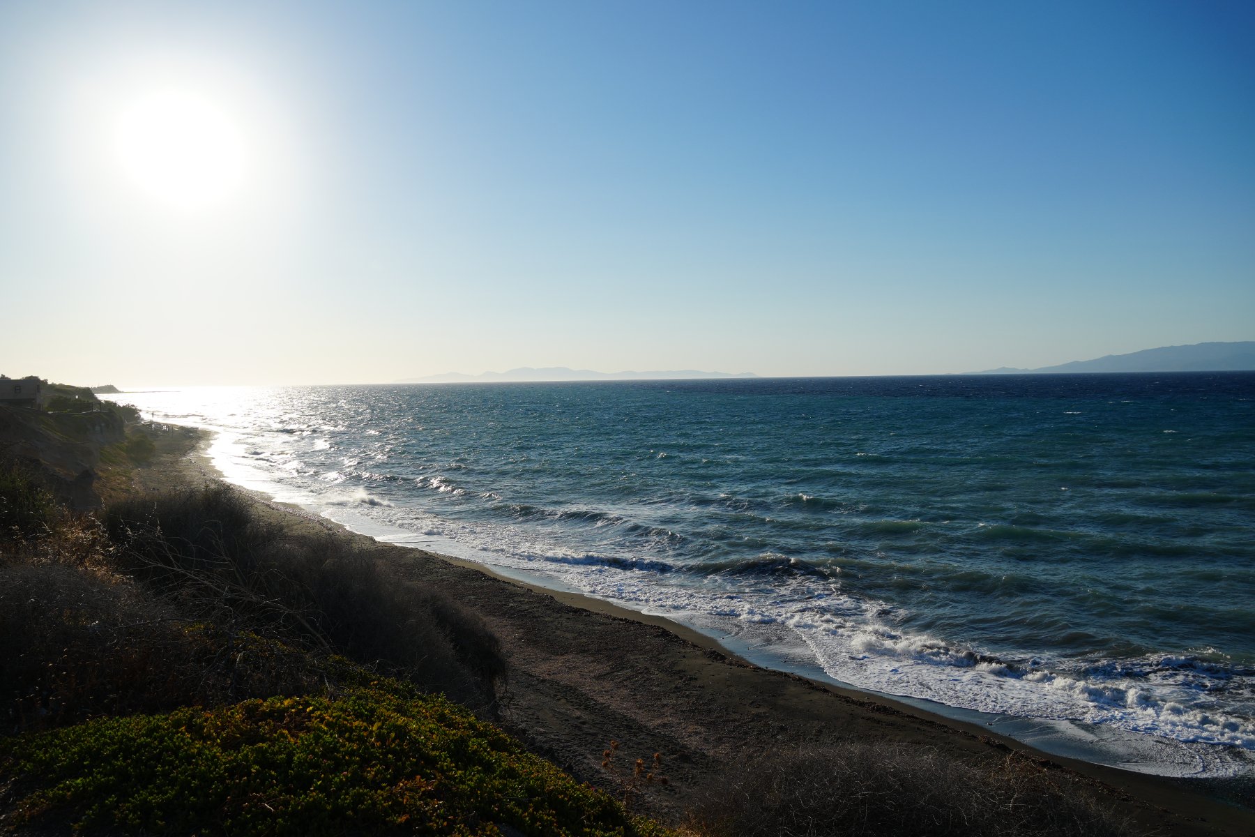 Late sun over a long curving beach, wind-whipped surf rolling in under a clear blue sky
