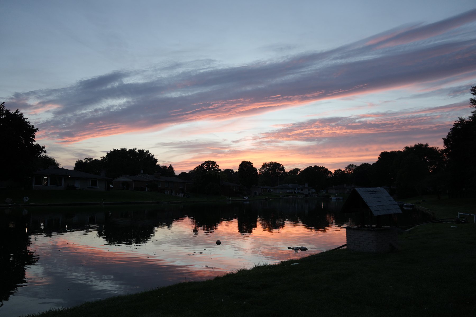 Pink and violet streaked clouds at dusk mirrored in a still lake lined with silhouetted trees and houses