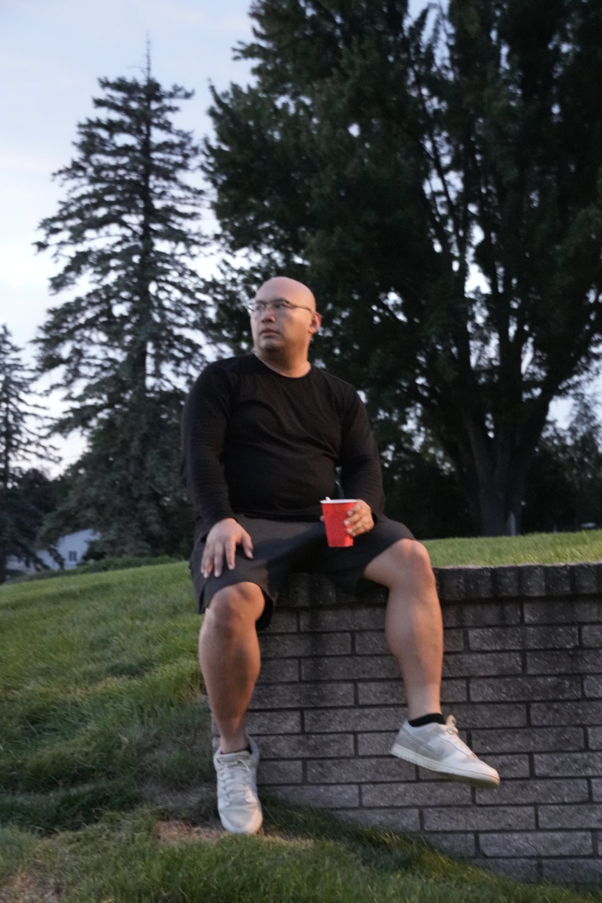 A man in black seated on a brick wall at dusk holding a red cup, tall pines behind