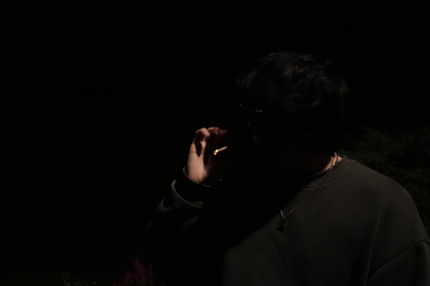 A figure in near-total darkness, the glowing ember at his fingertips the only light