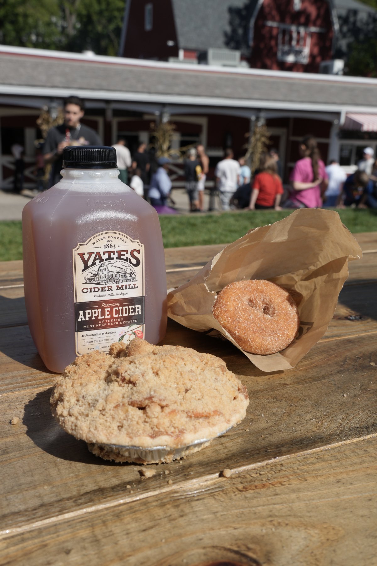 A jug of Yates apple cider beside a crumb-topped apple pie and a sugared cider donut on a wooden picnic table