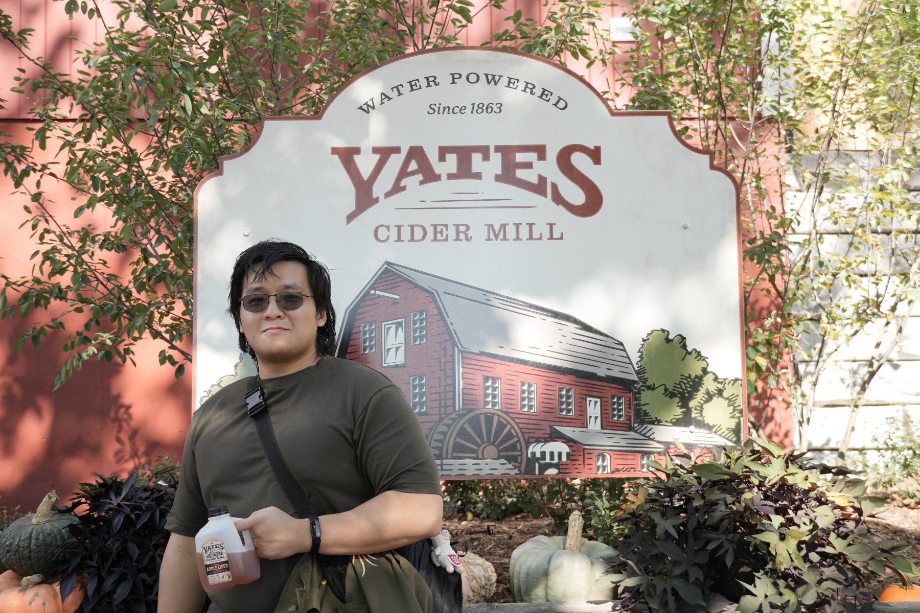 A man in sunglasses holding a jug of cider before the Yates Cider Mill sign, pumpkins at his feet