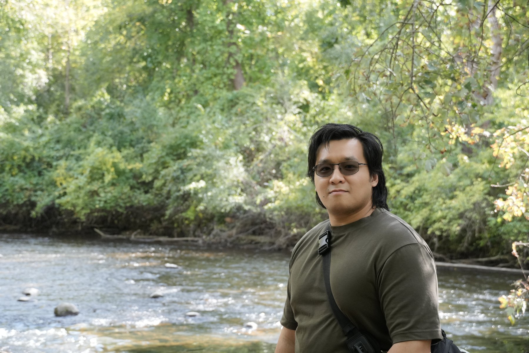 Portrait of a man in tinted glasses and an olive tee beside a sunlit river