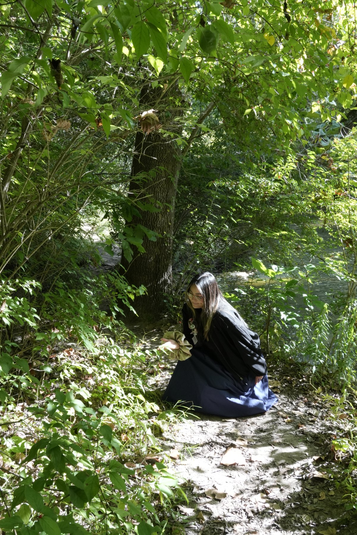 A woman kneeling on a shaded forest path beneath a canopy of green leaves, a river glinting behind
