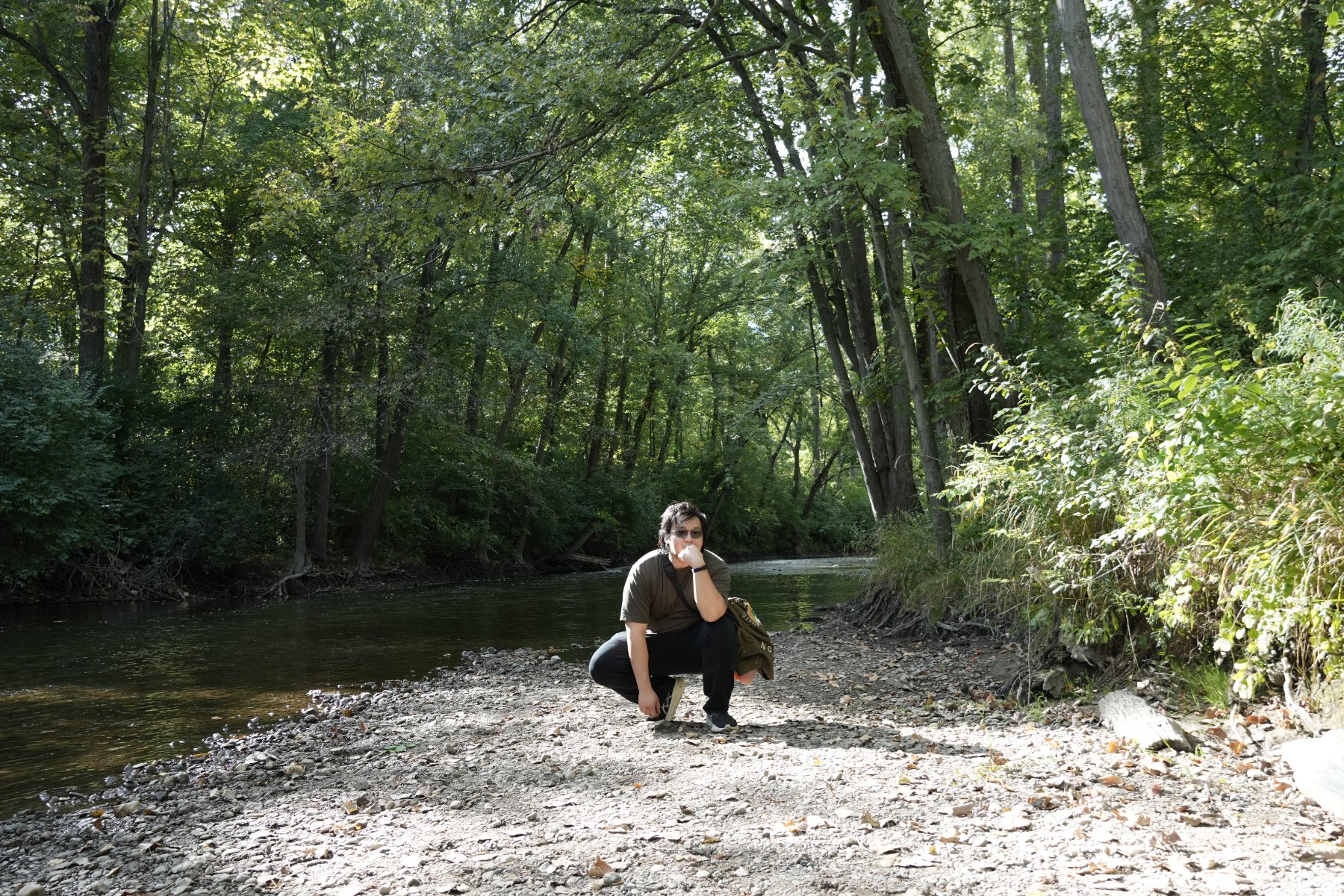 A man crouching thoughtfully on a gravel riverbank beneath an arch of trees
