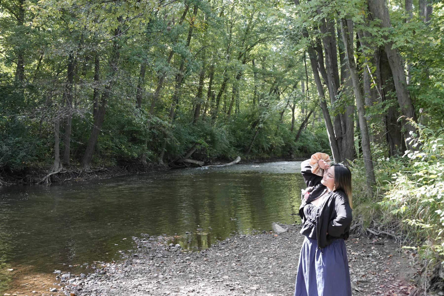 A woman shading her eyes on a pebbled riverbank under tall trees