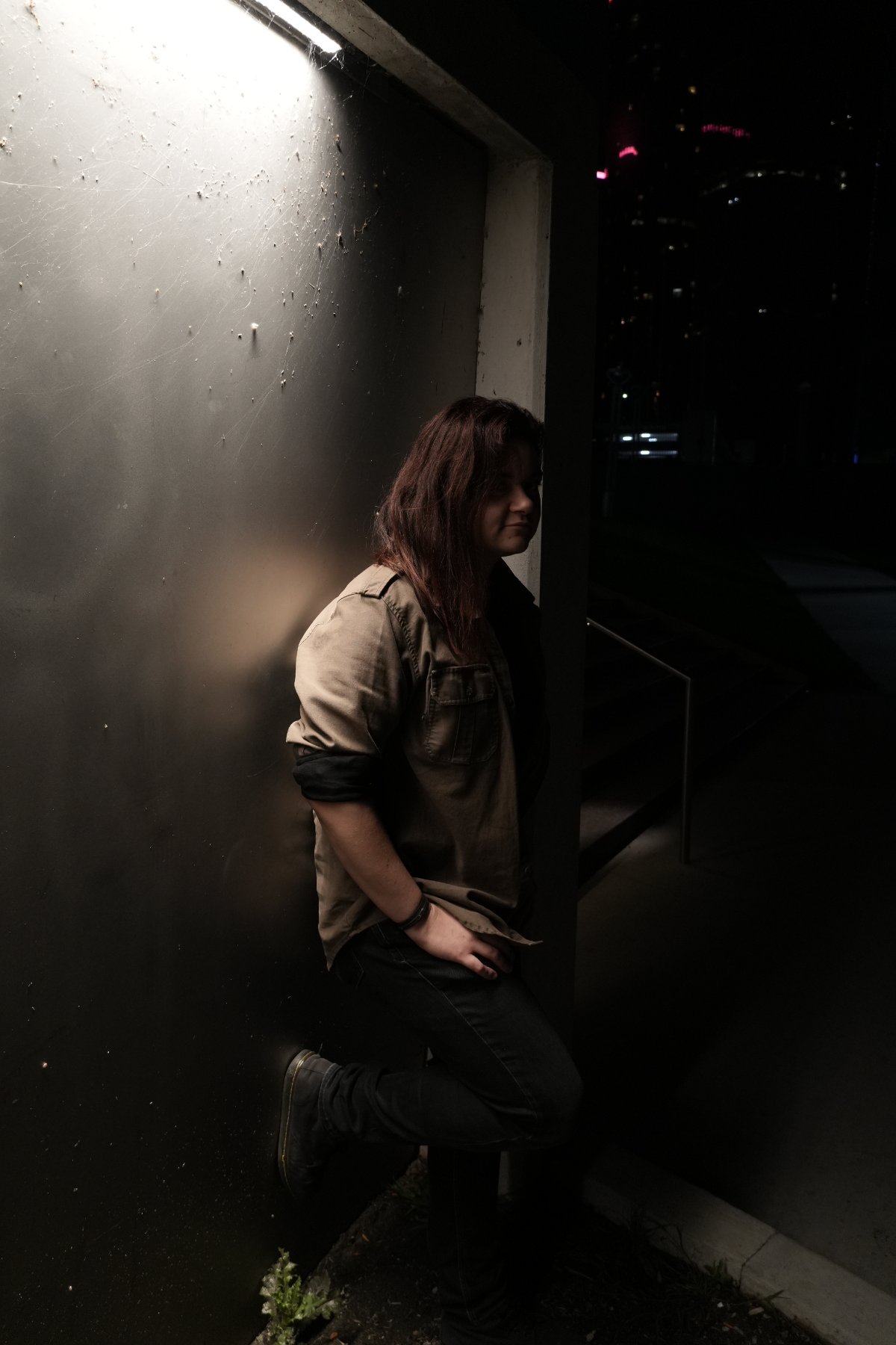 A figure in an olive shirt leaning against a dark wall beneath a harsh overhead light, city towers glowing behind
