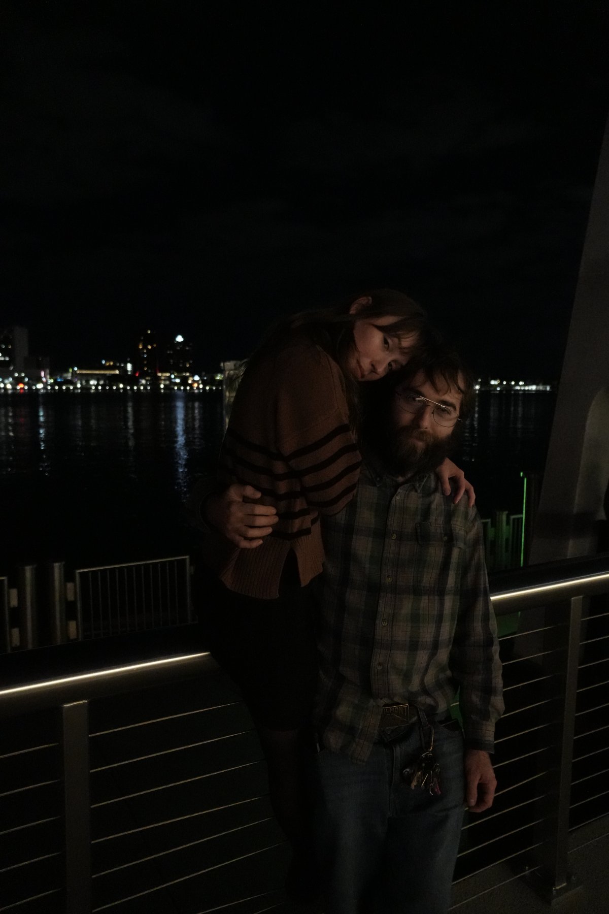 A woman in a striped sweater draped over a bearded man in plaid at a riverfront railing, city lights across the water