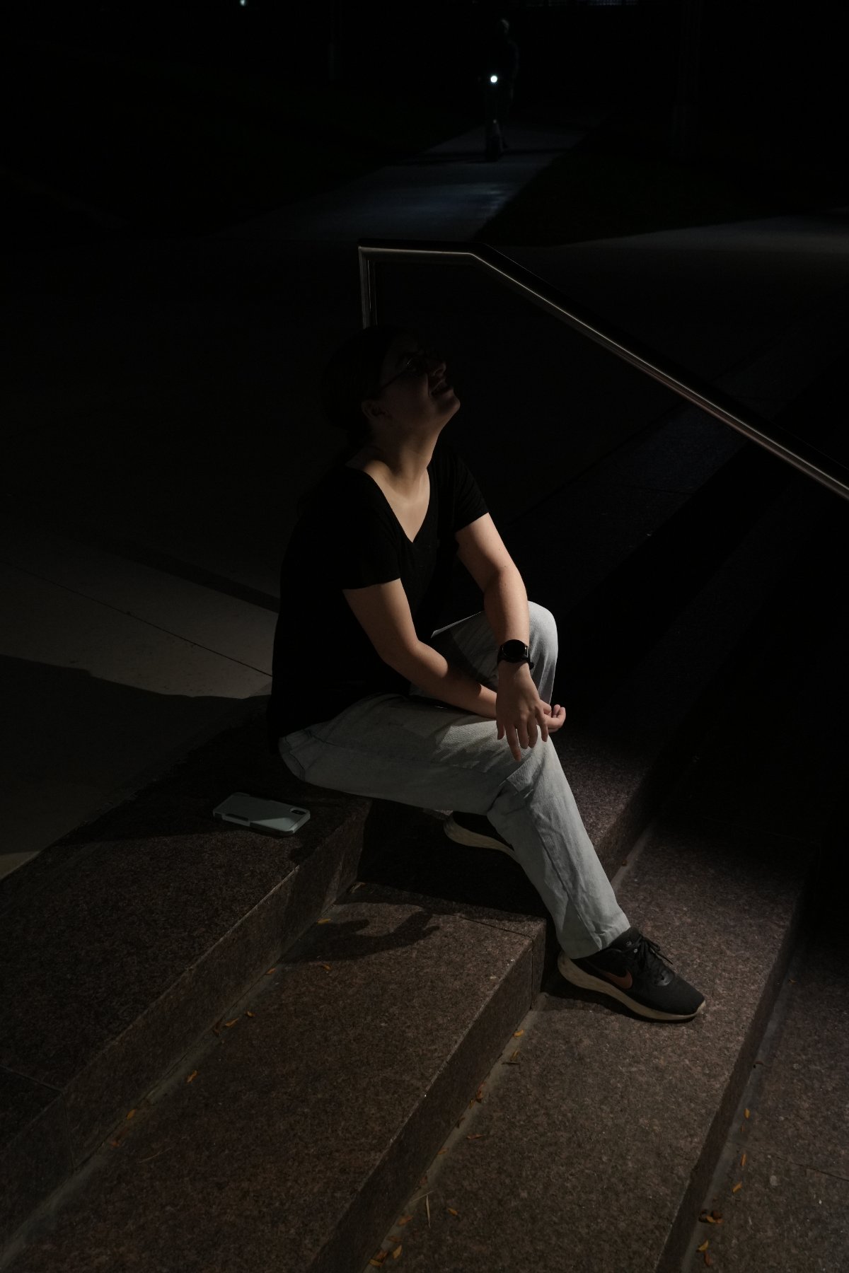A figure seated on granite steps at night, face tilted up into a single pool of light beside a steel handrail