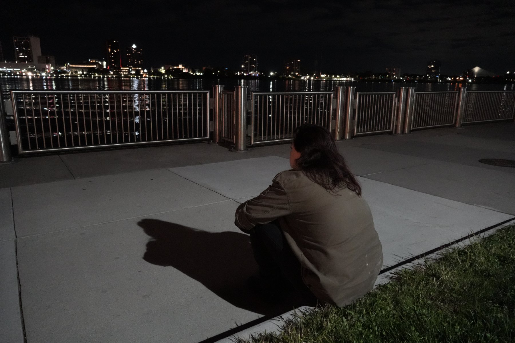 A figure seen from behind, sitting on the riverwalk facing a lit skyline across the water, a long shadow cast forward