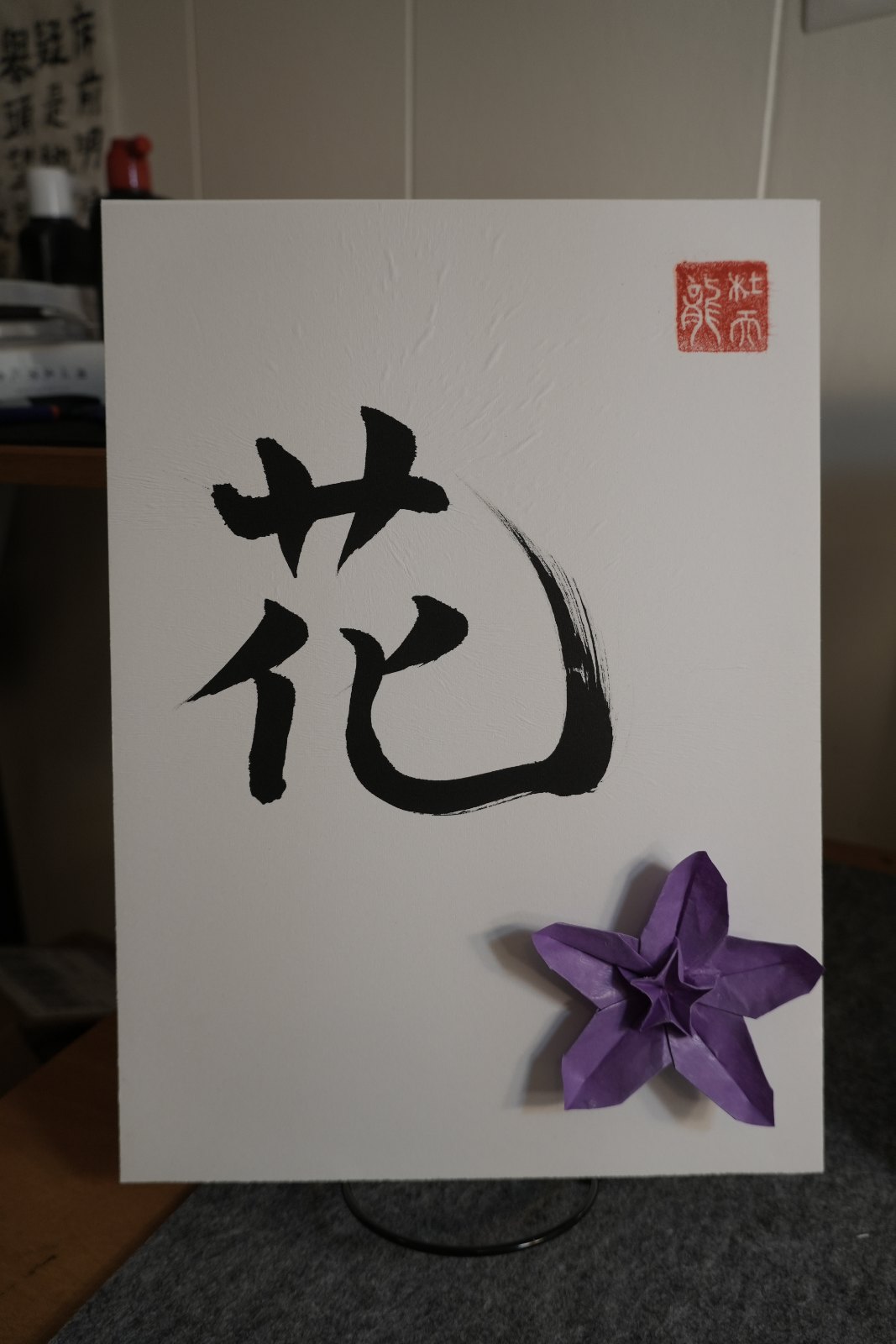 Shodo calligraphy of the kanji 花 (hana, flower) in black ink with a red seal, paired with a purple origami flower