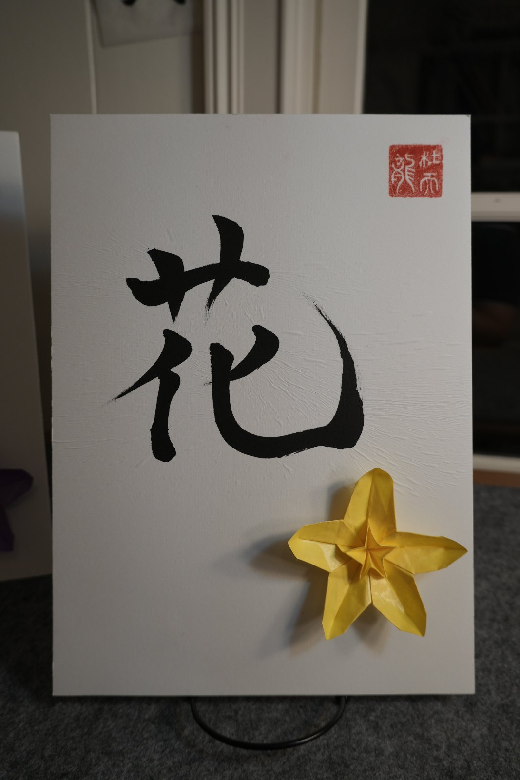 Shodo calligraphy of the kanji 花 (hana, flower) in black ink with a red seal, paired with a yellow origami flower