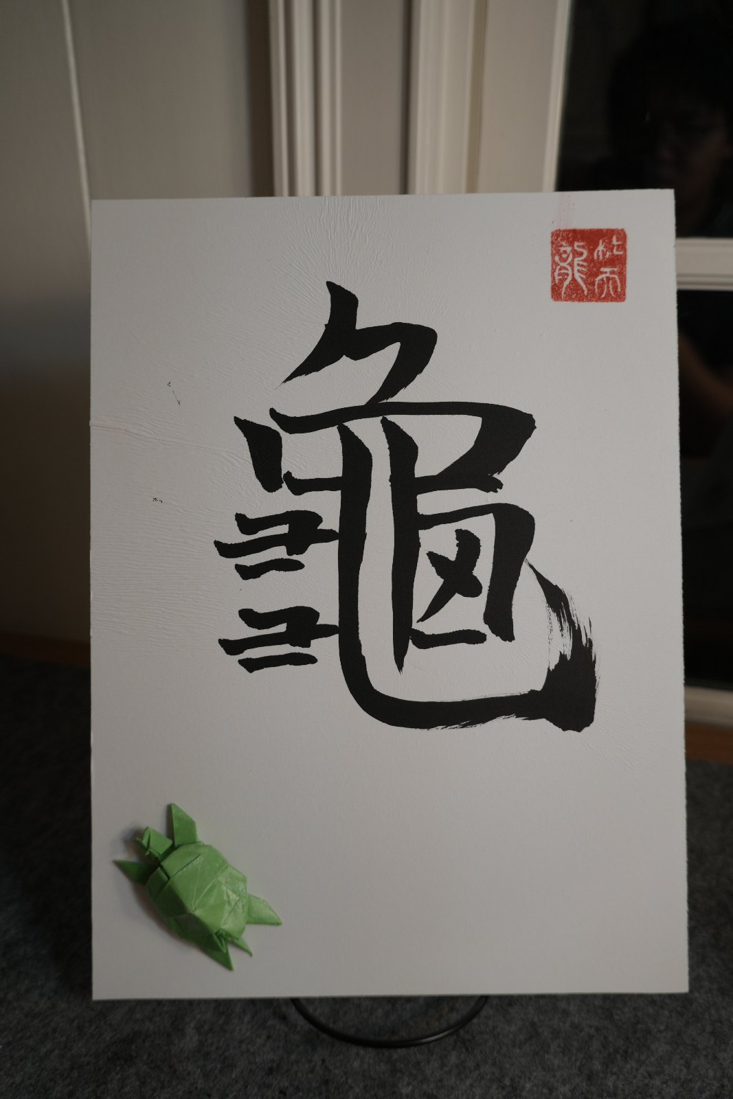 Shodo calligraphy of the kanji 亀 (kame, turtle) in bold black brushwork with a red seal, paired with a green origami turtle