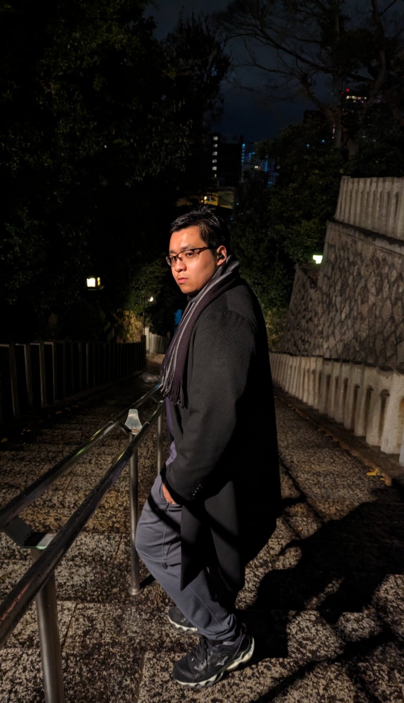 Skyler Đỗ in a dark wool coat and scarf, leaning on the railing of a stone stairway at night